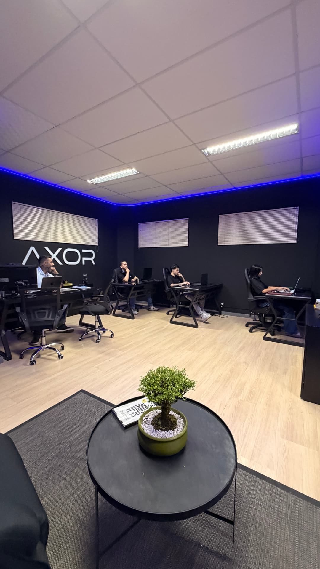 Sala de trabalho da AXOR, com a equipe em suas estações e o letreiro da marca na parede.